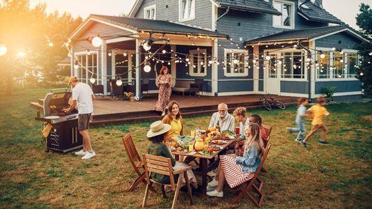 A family getting together for a day of grilling out outside of their house, while two children are playing off to the side.
